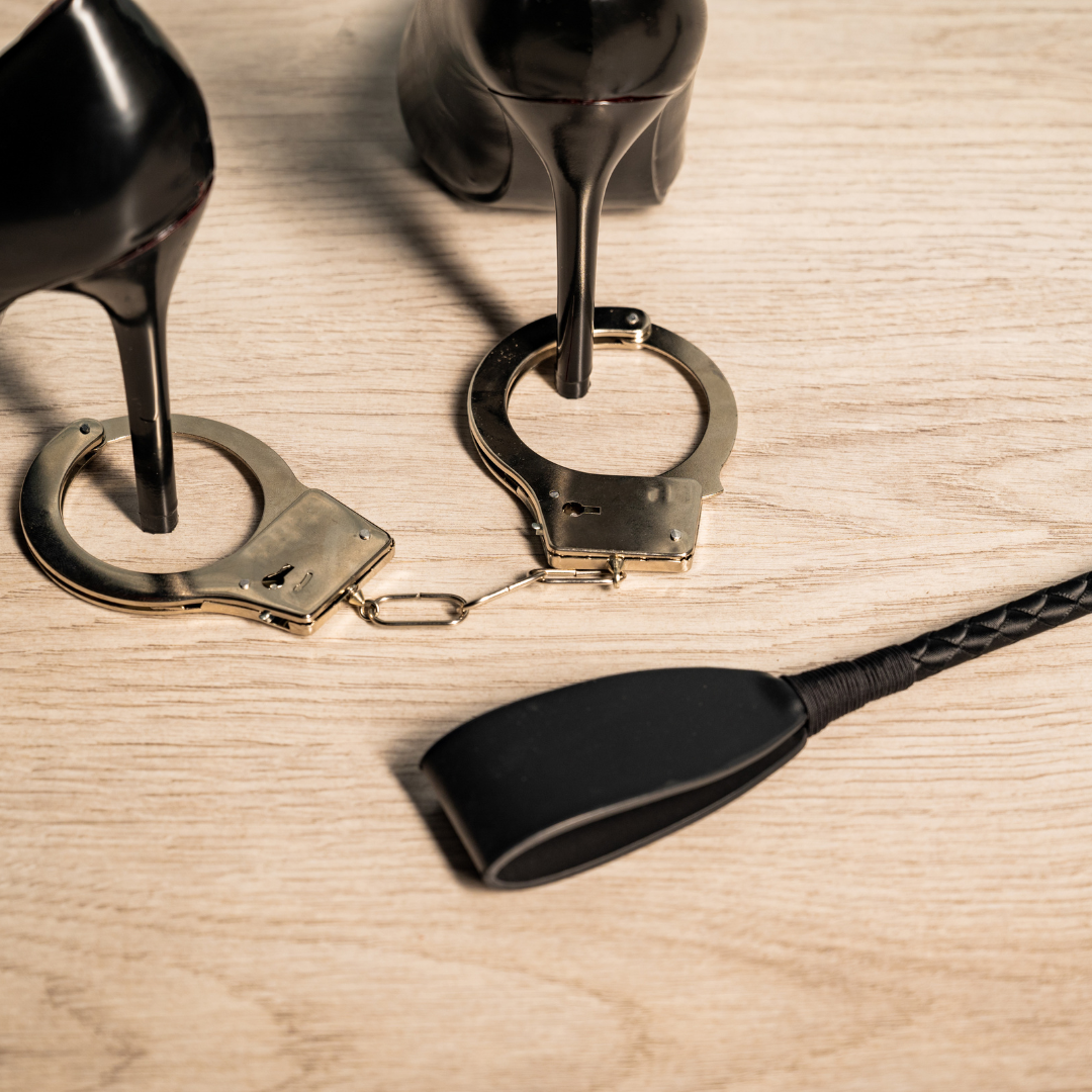 Black high heels, handcuffs, and a leather flogger on a wooden floor - BDSM accessories for kink play