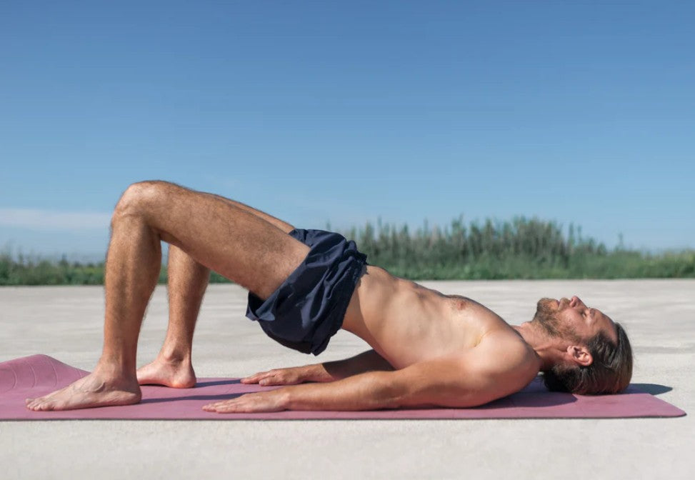 Man lying on a yoga mat outdoors in a bridge pose, lifting his hips to engage core and pelvic muscles.