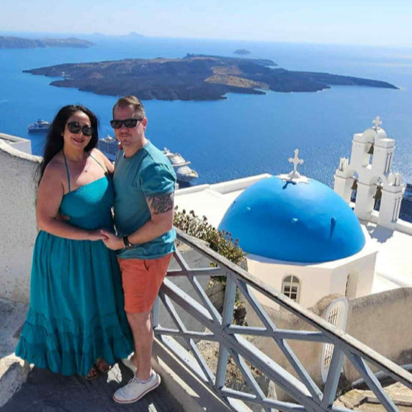 Linda Koontz and husband posing in front of a blue-domed church in Santorini Greece with a scenic view of the ocean and another island.