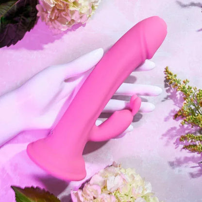 Close-up of Somebunny to Love in a hand showing the textured shaft and powerful rabbit ears in creamy silicone with suction base on background with flowers.