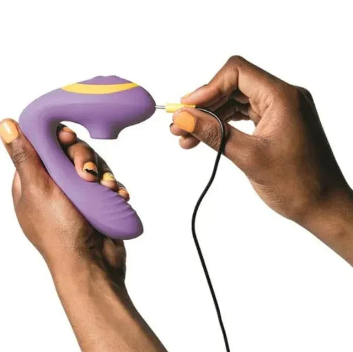 Purple ROMP Reverb dual stimulation vibrator being held in one hand while the charging cable is plugged in, showing the device's ergonomic shape and wireless charging capability.