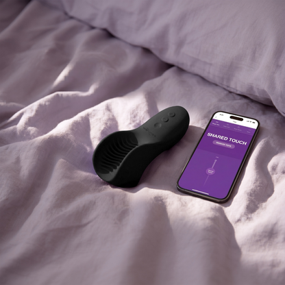 Black We-Vibe Shared Touch vibrator beside a smartphone displaying the We-Vibe app controls.